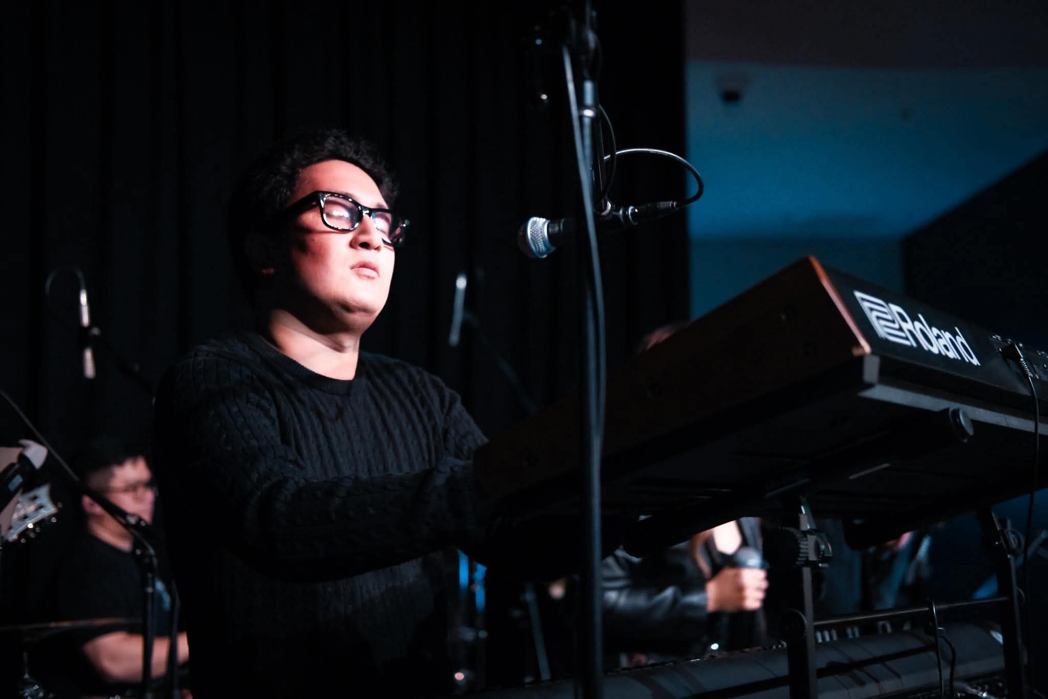 Claro wearing a black textured sweater and dark glasses, playing a Roland electronic keyboard on a dimly lit stage with blue stage lighting at West HQ with the Mayora's Project Band.